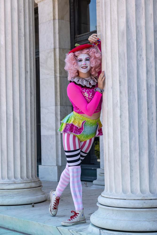 A colorful clown peeks from behind a column outdoors.