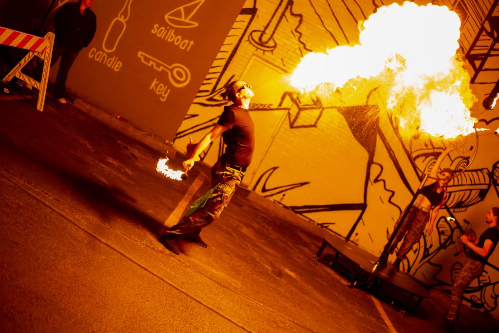 A person breathing fire against a graffiti wall at night.