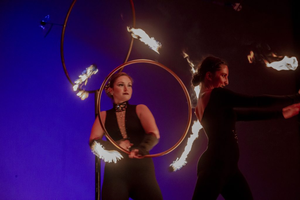 Two performers with flaming hoops in a dark setting.