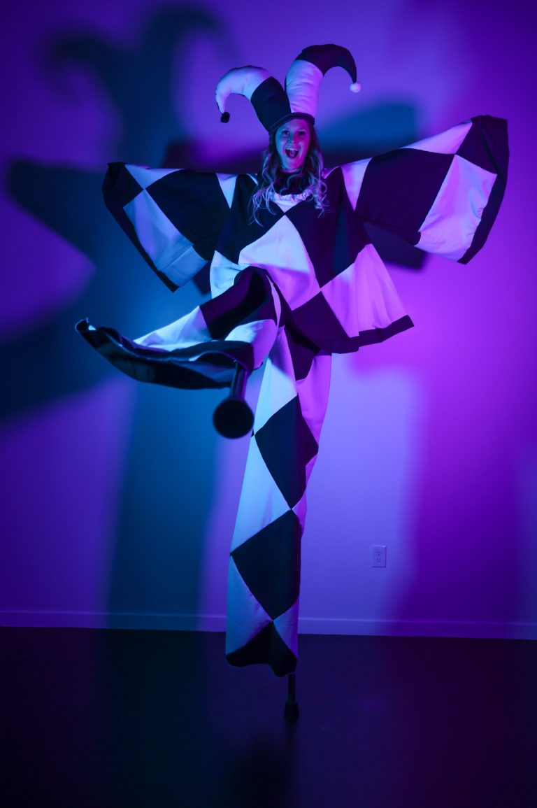 Person in harlequin costume kicking against purple-lit background.