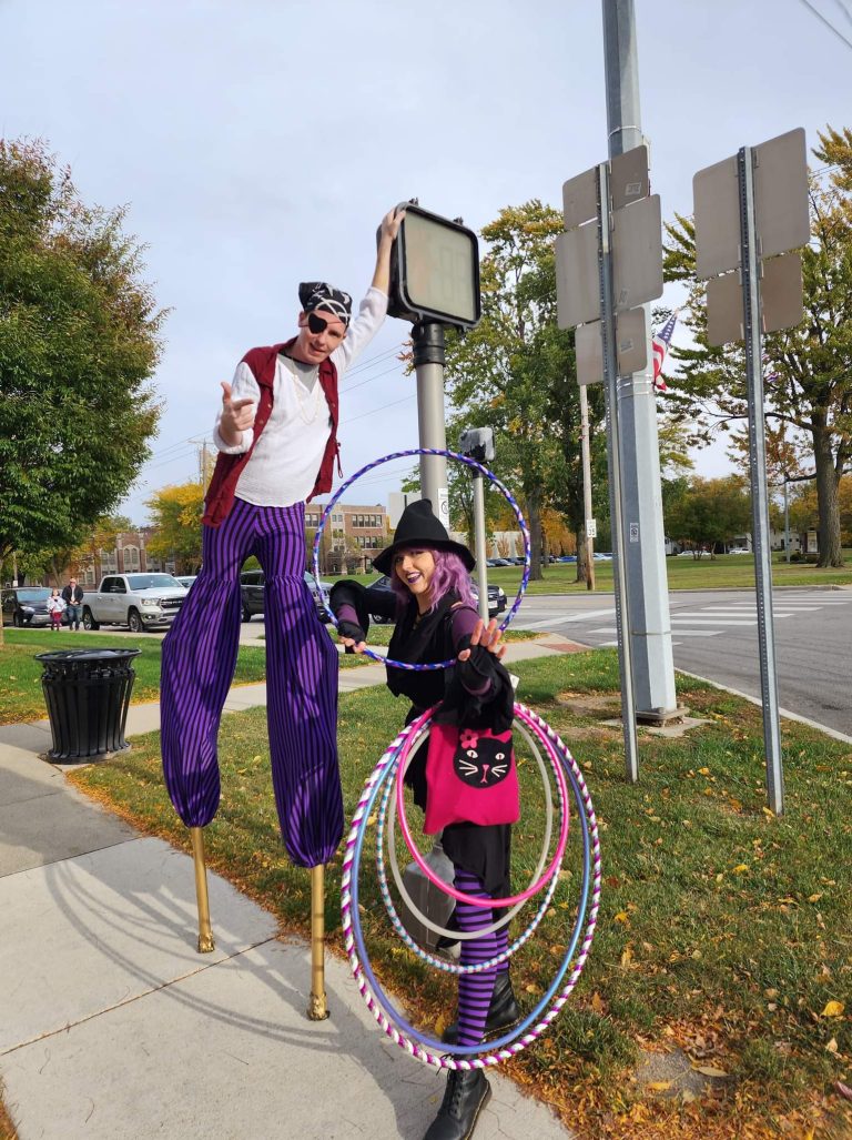 Two performers, one on stilts and one with hula hoops, entertain outdoors.