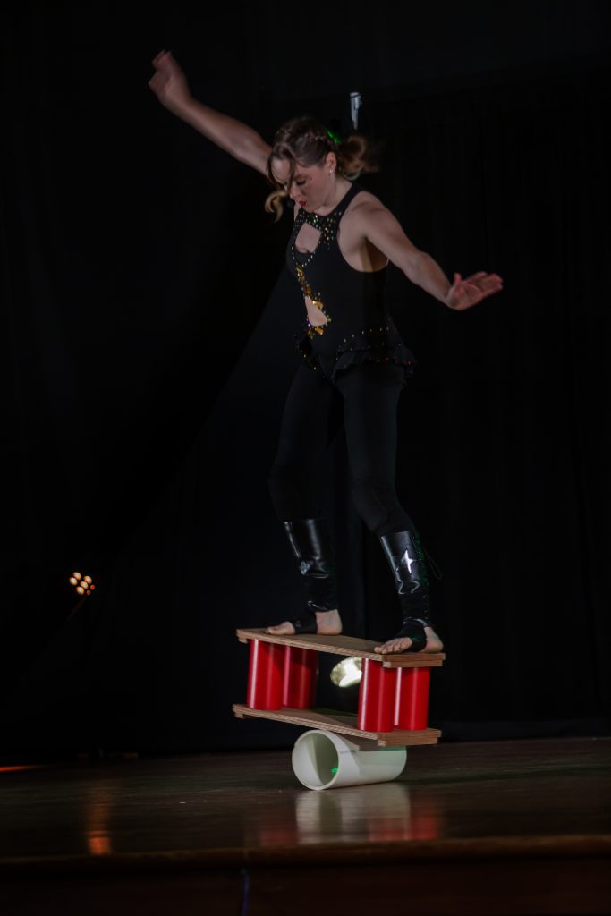 A performer balances on a chair stack on stage against a black background.