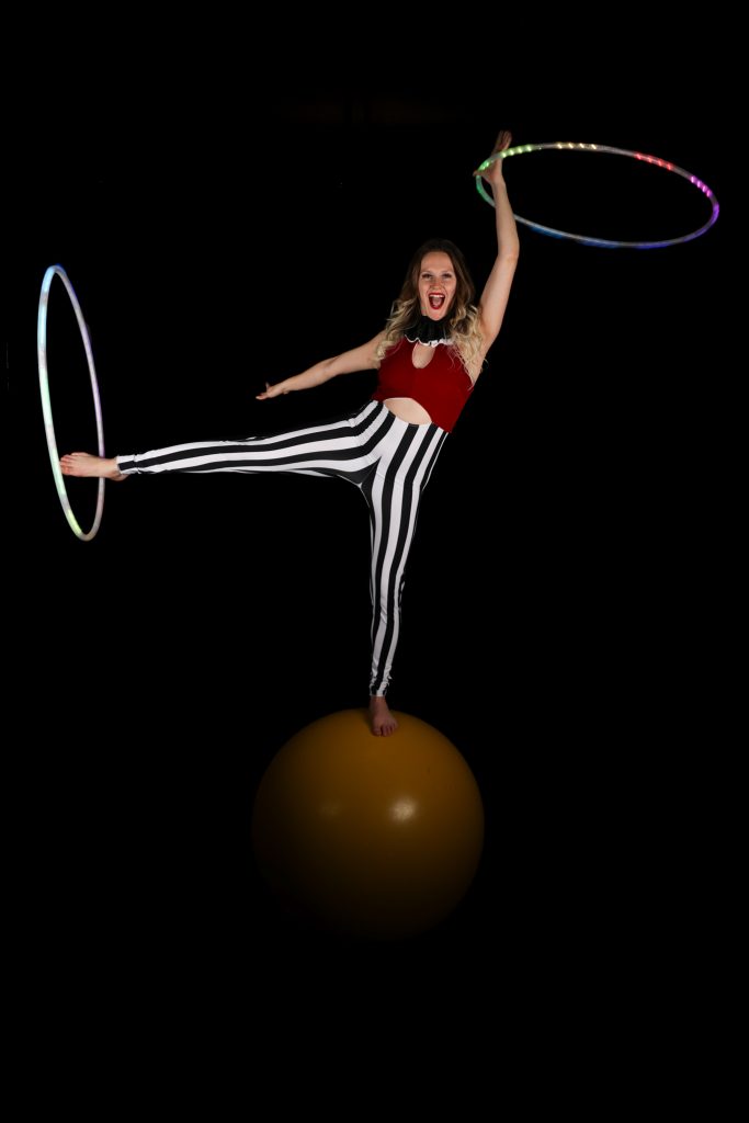 A circus performer balances on a large ball with a hula hoop.