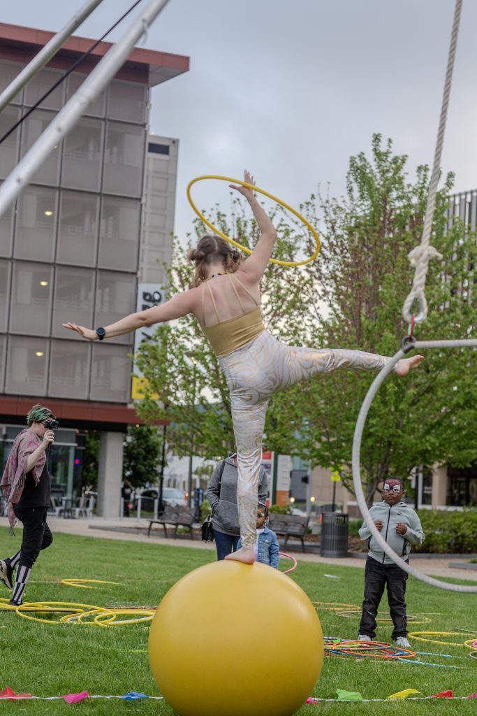 A performer balances on a ball while holding a hoop in an outdoor setting.