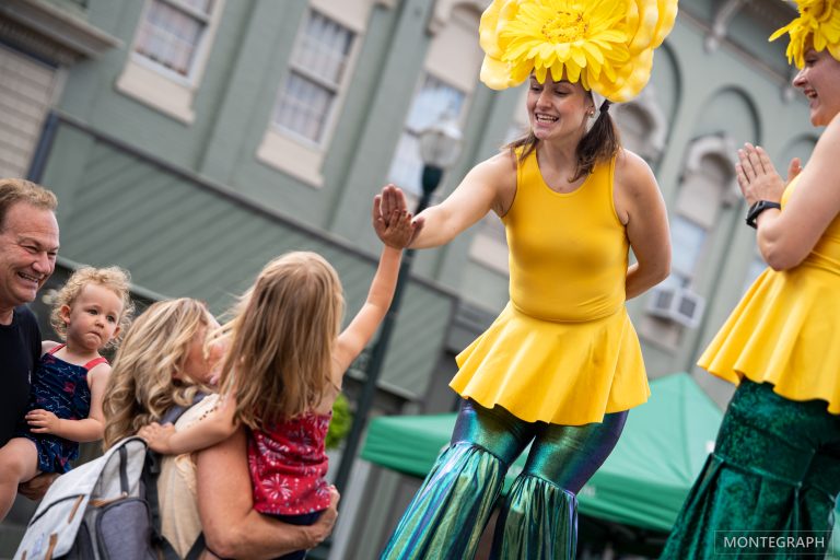 A person wearing a yellow top, yellow hat, and green striped pants holding hands with two children.