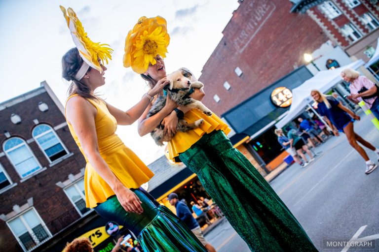 Two performers on stilts wearing colorful costumes with yellow feather fans.