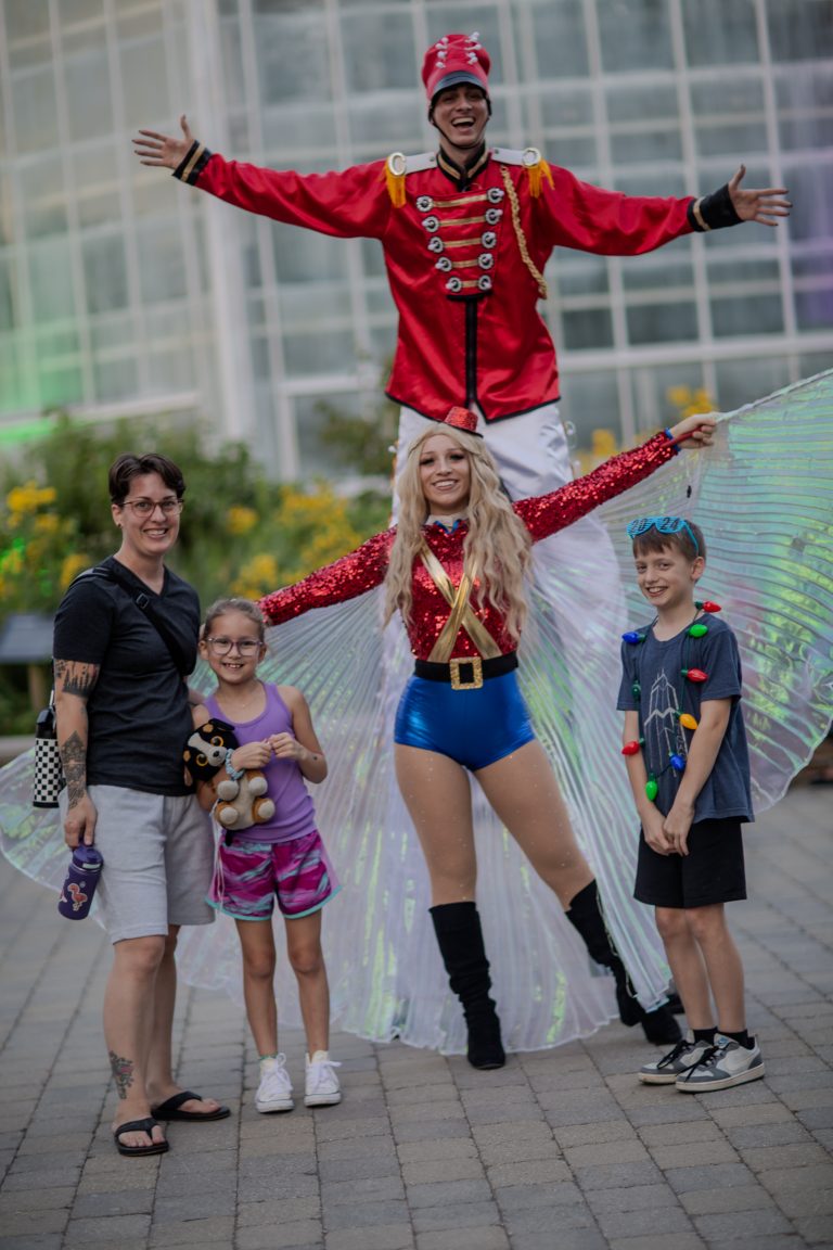 A woman performer on stilts posing with three children outdoors.