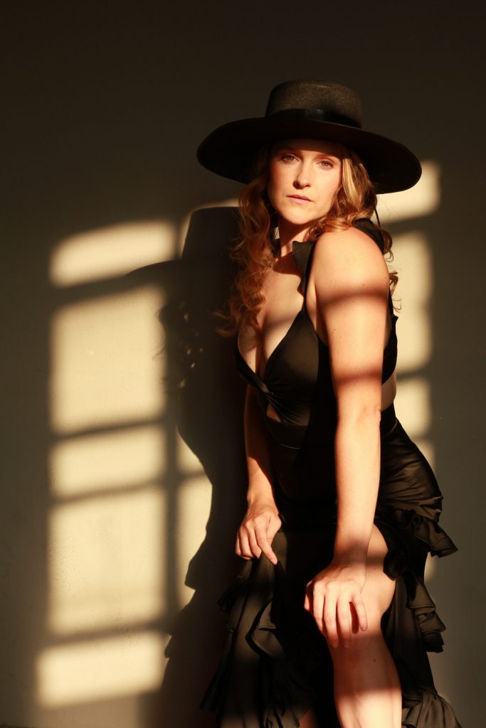 A woman in a black dress and hat posing with shadow patterns on her face.