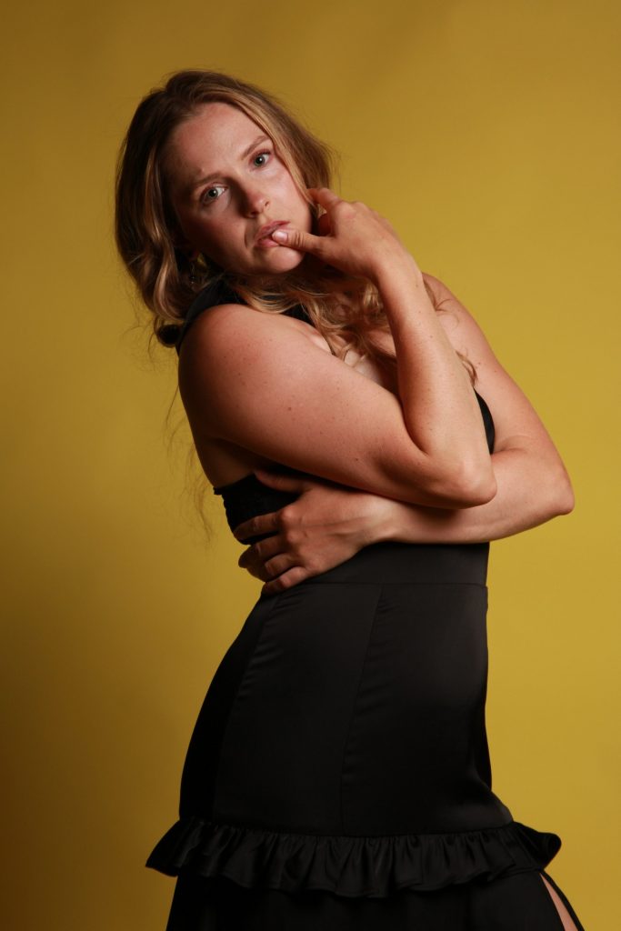 A woman posing against a yellow background in a black dress.