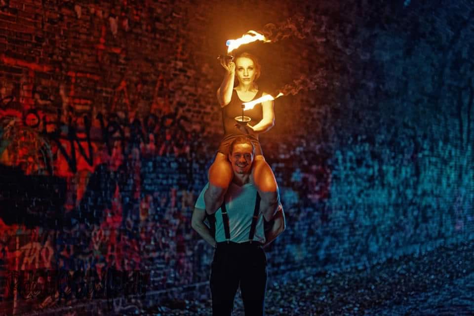 A man carrying a fire performer on his shoulders in a dark, graffiti-covered alley.
