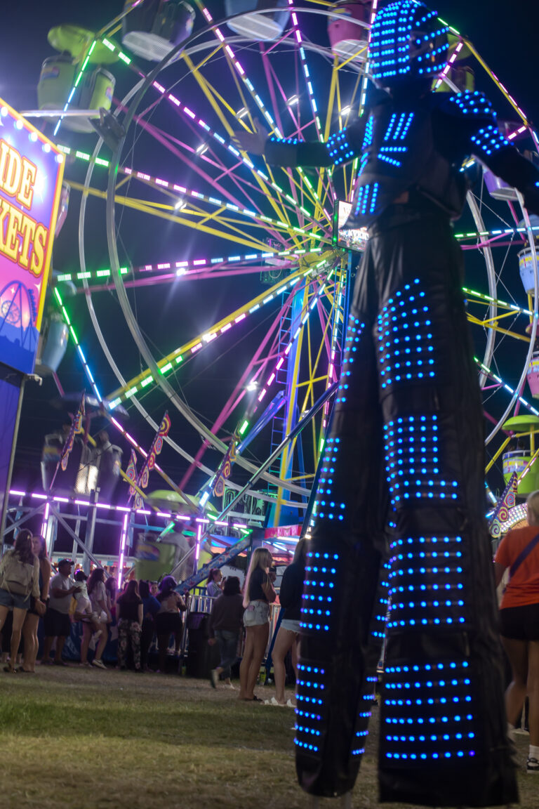 Stilt performer near illuminated Ferris wheel at night.